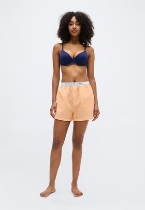 Žena s kovrčavom kosom stoji bosa, nosi mornarsko plavi grudnjak i narančaste prugaste kratke hlače s bijelim pojasom Polo Ralph Lauren.