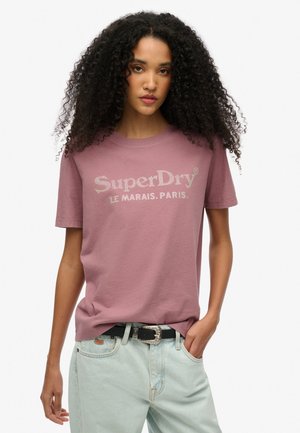 T-shirt en coton violet clair à manches courtes, avec un logo "SuperDry" pailleté et le texte "LE MARAIS. PARIS." sur le devant.