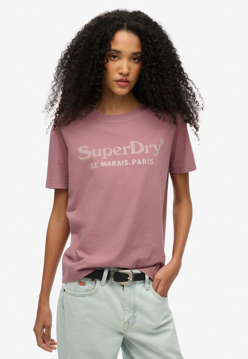 Camiseta de algodón de color lila claro con mangas cortas, que presenta un logo brillante de "SuperDry" y el texto "LE MARAIS. PARIS." en el frente.