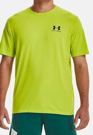 Mann trägt ein leuchtend gelb-grünes Under Armour kurzärmeliges T-Shirt und dunkelgrüne Shorts, steht mit den Armen an den Seiten vor weißem Hintergrund.