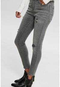 Mujer con jeans skinny rasgados grises, botas de tobillo negras con tachuelas y un suéter blanco, con una mano en el bolsillo.