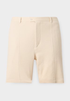 Jeune homme debout les mains dans les poches portant un t-shirt taupe, un short crème, des chaussettes blanches et des baskets beige sur un fond uni.