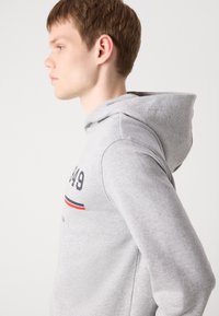 Sweat à capuche gris en tissu doux, doté d'une poche kangourou, d'une capuche avec cordon de serrage et d'une impression graphique à l'avant avec des accents rouges et bleus.