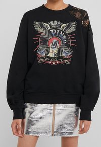 Felpa nera con design grafico colorato che presenta un microfono, ali e uccelli. Dettaglio in pizzo sulla spalla sinistra. Gonna riflettente argento sottostante.