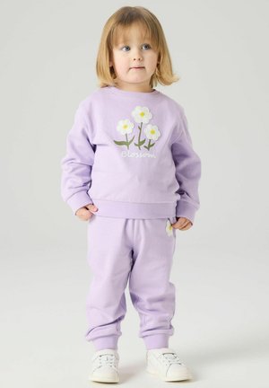 Bambino con capelli castano chiaro che indossa una tuta color lavanda con fiori bianchi e la scritta "Blossom", in piedi su uno sfondo chiaro uniforme.