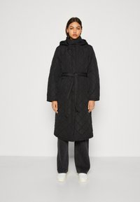Cappotto lungo trapuntato nero con cappuccio, vita regolabile con cintura e tasche laterali, caratterizzato da un motivo a rombi e una vestibilità comoda. Abbinato a pantaloni scuri.