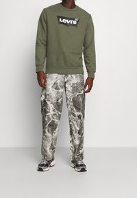 Sweat-shirt vert olive avec logo noir, associé à un pantalon à motif marbré gris. Modèle portant des baskets noires et grises, se tenant sur un fond uni.