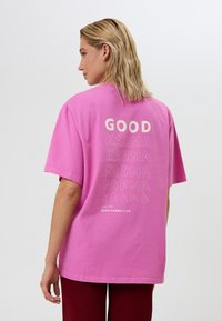 Camiseta de algodón rosa con un corte holgado, que cuenta con "BUENA KARMA" en varios estilos tipográficos en la parte posterior. Mangas cortas, escote redondo.