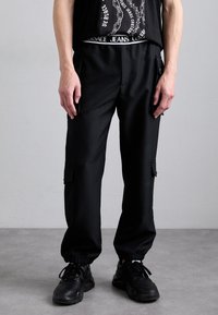 Calças cargo pretas com punhos elásticos, múltiplos bolsos com zíper e uma textura lisa e sólida. Usadas com uma t-shirt gráfica preta e ténis pretos.