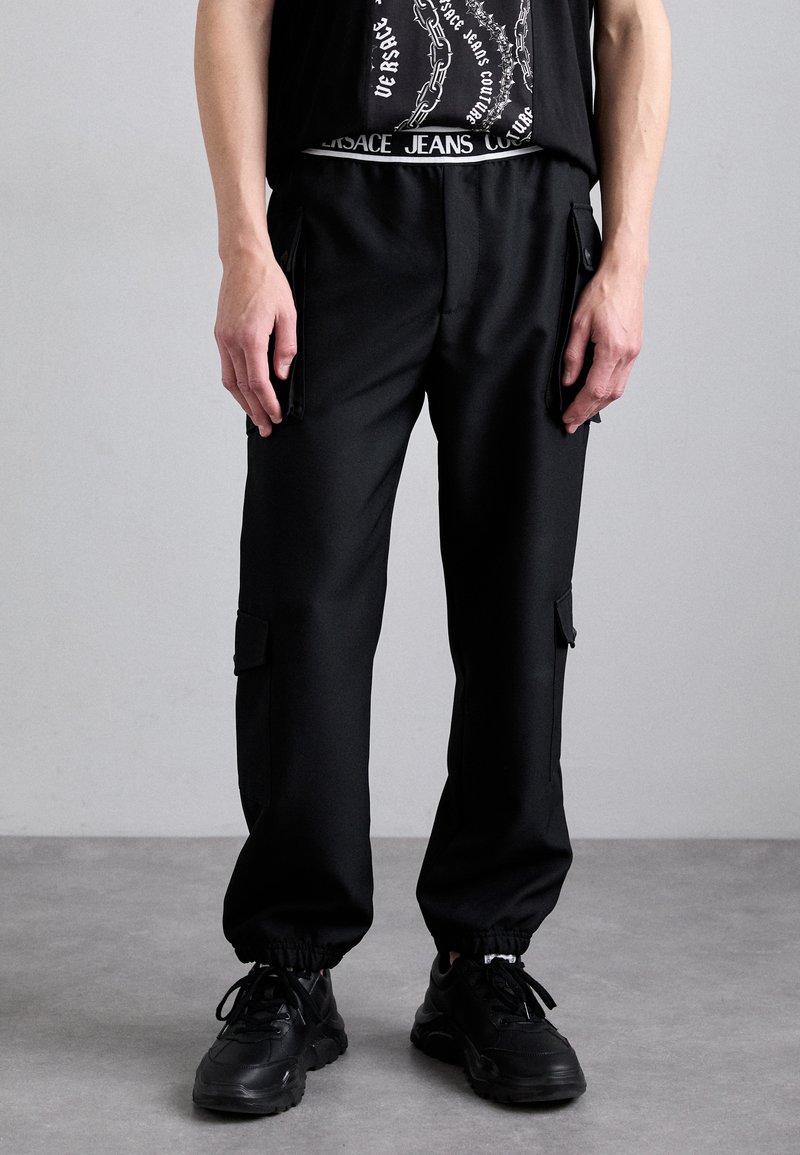 Calças cargo pretas com punhos elásticos, múltiplos bolsos com zíper e uma textura lisa e sólida. Usadas com uma t-shirt gráfica preta e ténis pretos.