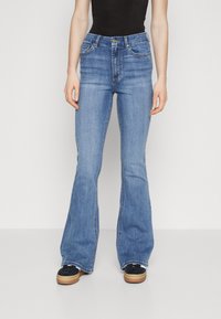 Ženske modre flare jeans hlače iz denima, z sprednjimi žepi, visokim pasom in kontrastnim šivanjem. Ujemajo se z črnimi čevlji.
