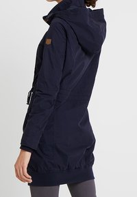 Granatowa, lekkia kurtka z kapturem, elastycznym pasem i mankietami; ma teksturowane wykończenie oraz małą naszywkę z logo na ramieniu.