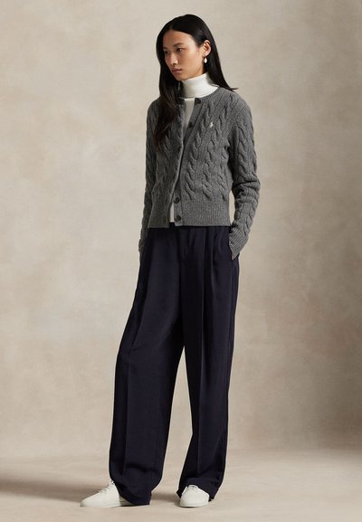 Nő szürke kábelkötött kardigánban fehér garbó felett, tengerészkék bő szárú nadrágban és fehér tornacipőben, kezét zsebében tartva áll.