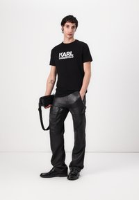 T-shirt en coton noir avec logo 'KARL LAGERFELD' blanc, associé à un pantalon en cuir noir avec poches et coupe slim. Chaussures et sac noirs.