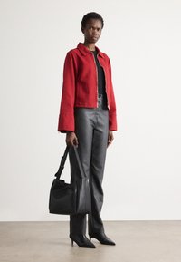 Kvinne med rød zip-up jakke, svart topp, svarte skinnbukser, spisse hæler, og som holder en svart skinnveske mot en ensfarget bakgrunn.