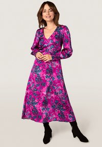 Robe florale rose avec des manches longues, un décolleté en V profond et une silhouette fluide, agrémentée d'accents bleus et violets. Des bottines noires à la cheville complètent le look.
