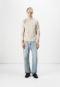 Maglione a collo alto in maglia a treccia di colore crema, abbinato a jeans a gamba larga di un blu chiaro. Il modello indossa scarpe nere. Lo sfondo semplice e pulito mette in risalto l'abbigliamento.