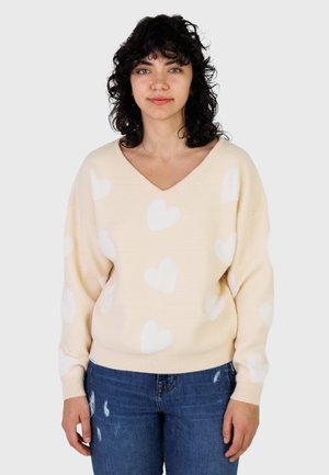 Jeune femme aux cheveux bouclés et foncés, portant un pull beige avec des motifs de cœurs blancs et un jean bleu déchiré, debout devant un fond uni.