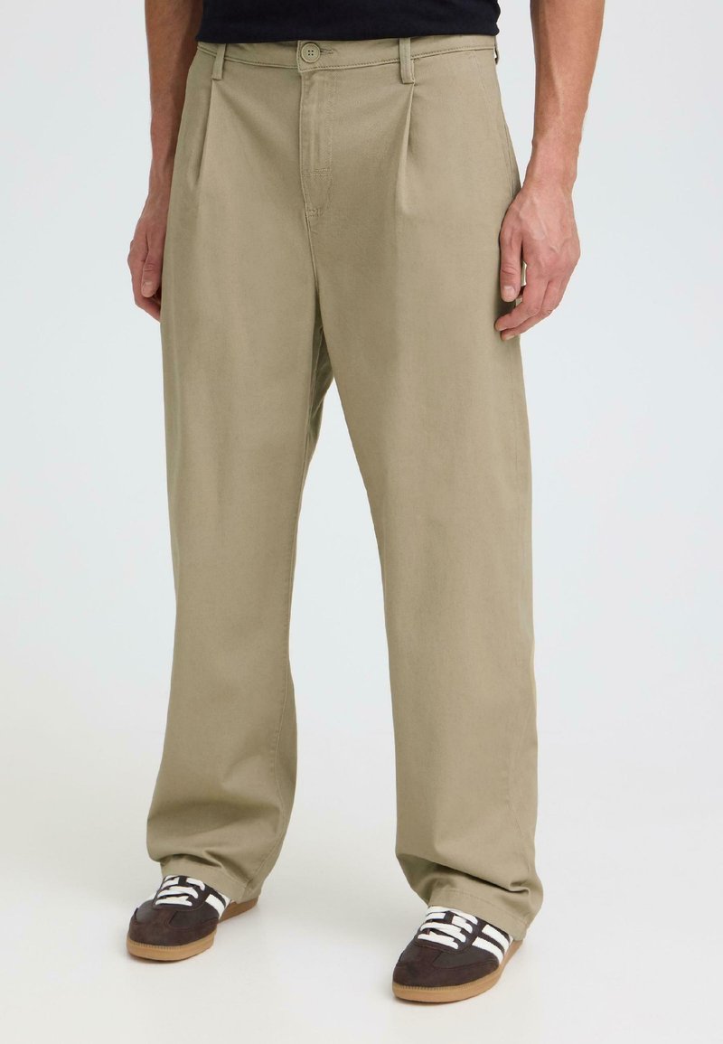 Homme portant un pantalon beige ample et des baskets noires à rayures blanches, debout devant un fond clair uni.