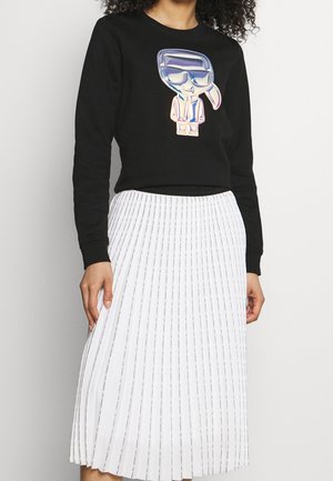 Person iført sort langærmet skjorte med skinnende tegneseriefigur og hvid plisseret midi-nederdel med tynde lodrette striber.