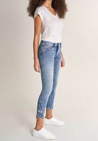 Světlé modré skinny džíny s vysokým pasem, s roztrhaným lemem a bočními ventilačními otvory, ve spojení s bílým přiléhavým tričkem a bílými teniskami.