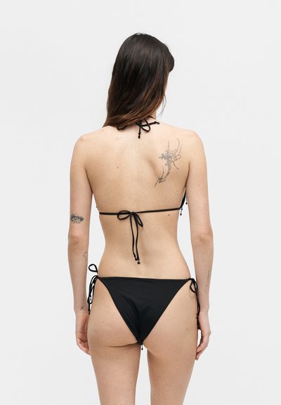 Γυναίκα με μαύρο στρινγκ μπικίνι, εμφανίζεται από πίσω, αποκαλύπτοντας τατουάζ στην άνω πλάτη και τον αριστερό βραχίονα, με φόντο ένα απλό ανοιχτό χρώμα.