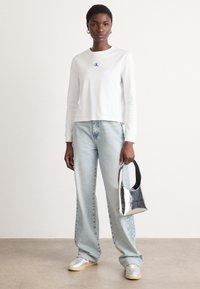 Camicia bianca a maniche lunghe con un piccolo logo blu, jeans dritti azzurri chiari, sneakers argentate e una borsa lucida argento con manico nero.