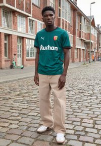 Grön fotbollsdräkt med svarta accenter, som har en logotyp och text. Beiga vida byxor och vita sneakers kompletterar outfiten. Stenlagd bakgrund.