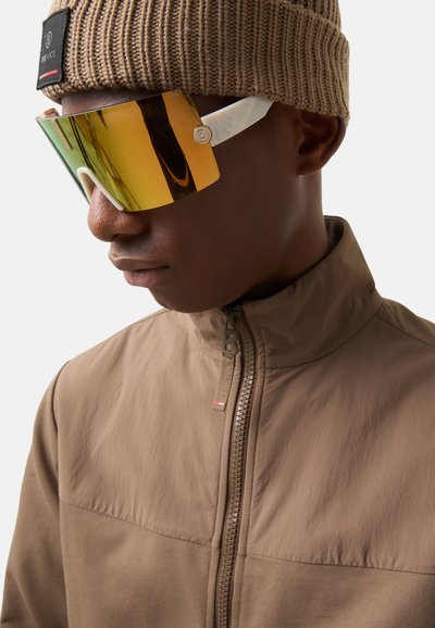 Sunglasses with a large amber visor and white frame, worn with a brown zip-up jacket and beige knit beanie. Smooth, reflective surface.