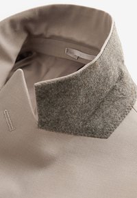 Gros plan sur le col d’un manteau beige avec un tissu en laine grise texturé sur le bord supérieur et des surpiqûres apparentes de boutonnière.