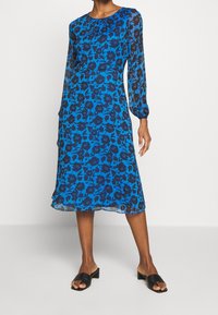 Kvinna klädd i en blå midikjol med svarta blommönster och långa genomskinliga ärmar, tillsammans med svarta öppna klackskor.