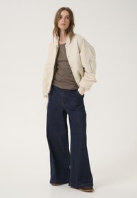 Veste bomber crèmée, débardeur marron, et jean foncé à jambe large. La veste dispose d'une poche zippée, d'une texture douce et de boutons-pression.