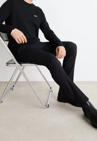 Pull noir à manches longues avec détails côtelés et logo, associé à un pantalon noir ajusté et des bottines noires, assis sur une chaise en métal.