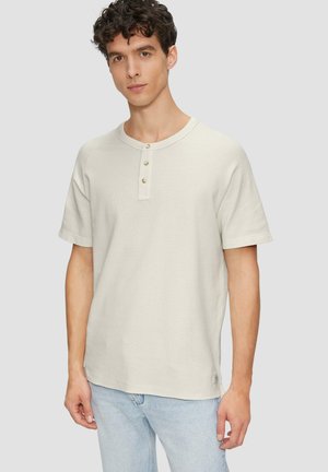 Camiseta de manga corta beige claro, fabricada con tela suave, que presenta un cuello henley con tres botones y un ajuste holgado.