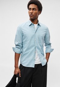 Ανοιχτό μπλε πουκάμισο με καρό σχέδιο και τσέπη, φορεμένο πάνω από ένα λευκό μπλουζάκι. Διαθέτει γυρισμένα μανίκια και καμπυλωτή τελείωση.