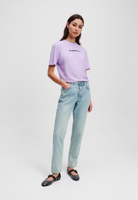 Lila katoenen T-shirt met korte mouwen, blauwe denim jeans en zwarte ballerina's met witte stippen; staande pose op een neutrale achtergrond.