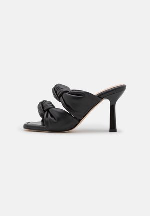 Mules de salto alto em couro preto, com tiras atadas, design de ponta aberta e salto stiletto. Textura suave com um acabamento semibrilhante.