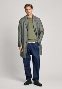 Abrigo de lana gris con solapa muesca, superpuesto a un suéter verde, combinado con pantalones de mezclilla azul y zapatos negros. Fondo sencillo.