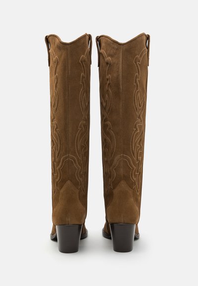 Brūnas zamšādas kovboju zābaki ar garām kājām, sarežģītām izšūtām detaļām, norakstītiem pirkstiem un bloku papēžiem. Divi vilkšanas cilpas augšpusē.