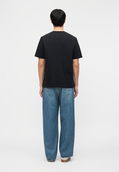 Άνδρας που στέκεται με την πλάτη γυρισμένη, φορώντας μια απλή μαύρη κοντομάνικη μπλούζα και φαρδιά μπλε τζιν με λευκά αθλητικά παπούτσια σε λευκό φόντο.