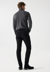 Sivi dugi rukav turtleneck top uparen s crnim uskim trapericama i crnim gležnjačama, prikazan sa stražnje strane na neutralnoj pozadini.