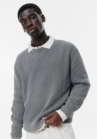 Maglione grigio a coste con colletto bianco, dotato di maniche lunghe e una vestibilità comoda. Il modello tiene l'orlo, mettendo in evidenza la texture e i dettagli del design.
