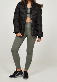 Chaqueta de plumas negra con capucha con borde de piel, cierre frontal con cremallera y detalles de logo, combinada con leggings acanalados verdes oliva y zapatillas negras.