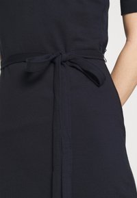 Dunkelblaues Kleid mit einem gebundenen Taillengürtel, das aus einem strukturierten Stoff gefertigt ist und kurze Ärmel hat. Die Nähte sind entlang der Seiten sichtbar.