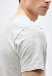 Homme portant un t-shirt à manches courtes gris clair, visible de l'épaule au menton, sur un fond blanc uni.