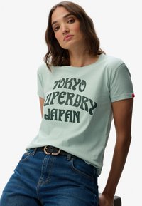 Lys grøn bomulds T-shirt med sort tekst, der siger "TOKYO SUPERDRY JAPAN." Den har en rund hals og korte ærmer, parret med blå jeans.