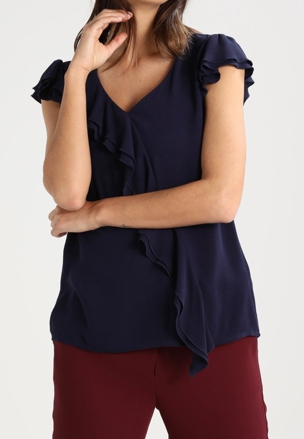 Femme portant un chemisier bleu marine à volants avec un col en V et des manches courtes bouffantes, ainsi qu'un pantalon bordeaux, bras croisés et main près du visage.