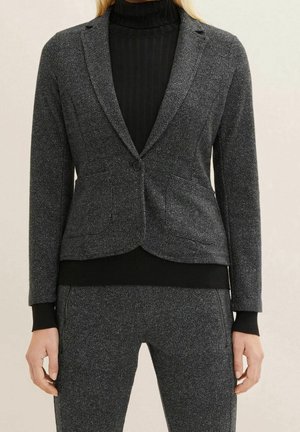 Kvinde iført en tætsiddende mørkegrå blazer og matchende bukser over en sort ribbet rullekravetrøje, stående mod en neutral baggrund.