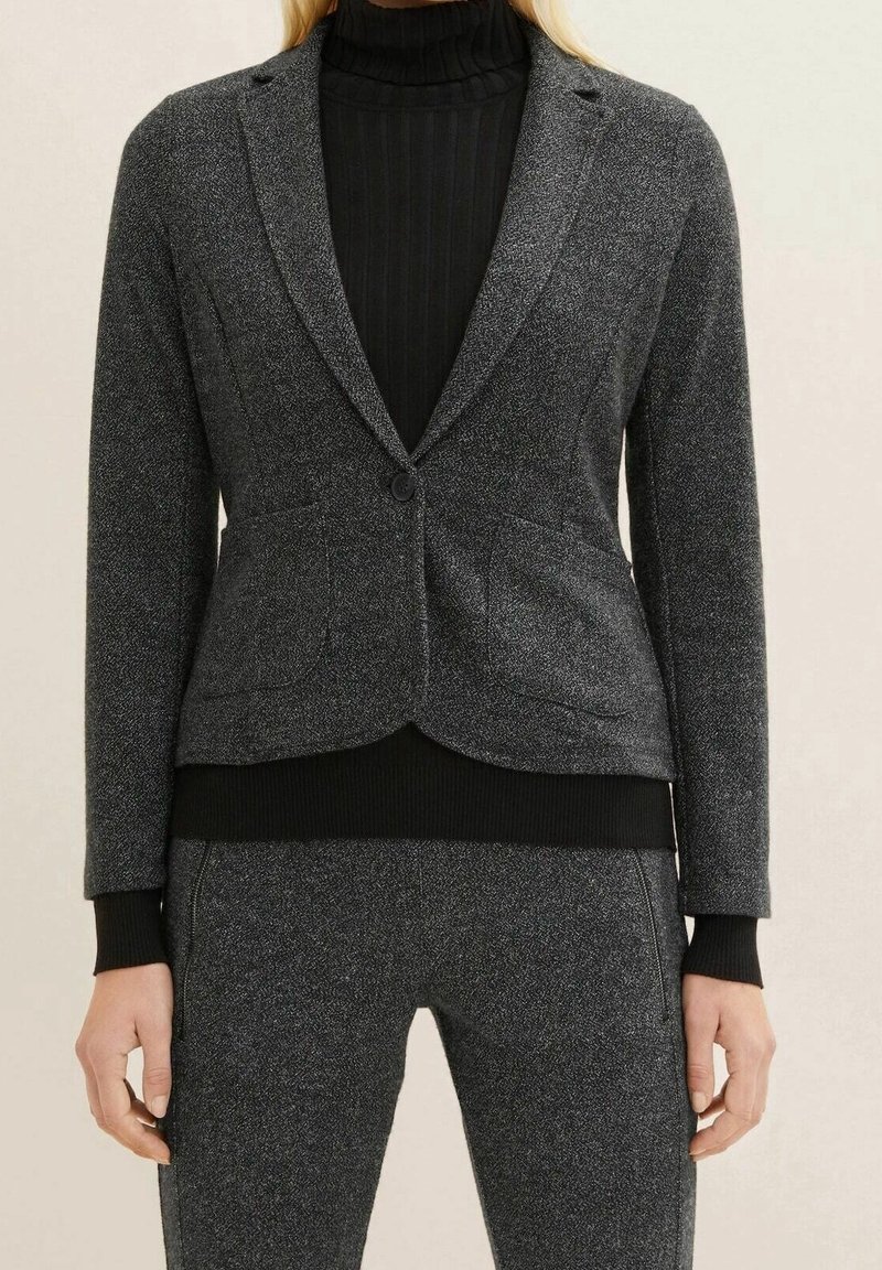 Femme portant un blazer gris foncé ajusté et un pantalon assorti sur un pull à col roulé noir côtelé, debout devant un fond neutre.