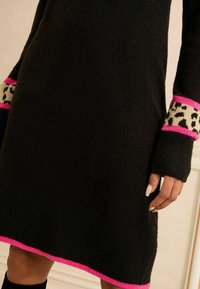 Abito in maglia nera con orlo rosa e dettagli in stampa leopardata sui polsini. Morbido al tatto, maniche lunghe e vestibilità leggermente ampia.